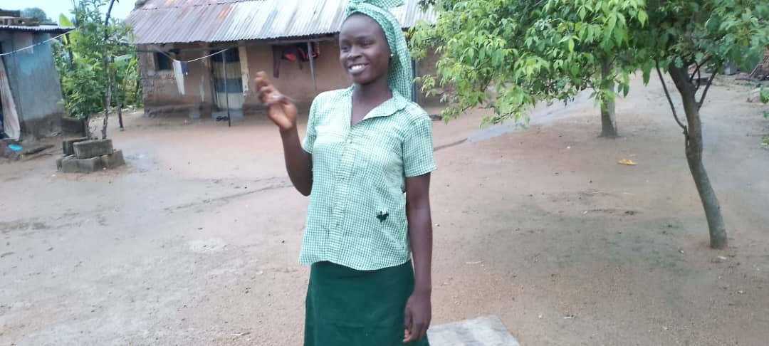 Agal stands outside her home smiling to the camera. She is in her school uniform.