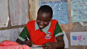 Violet sits at her desk writing. She is wearing her school uniform. her school fees are being supported by the project