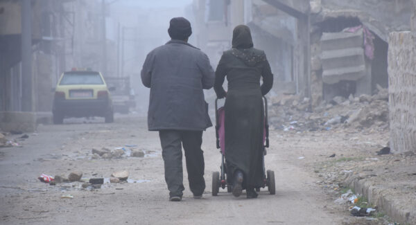 Parents walk their baby in a stroller in Aleppo. It is very cold and buildings around them are broken and destroyed.