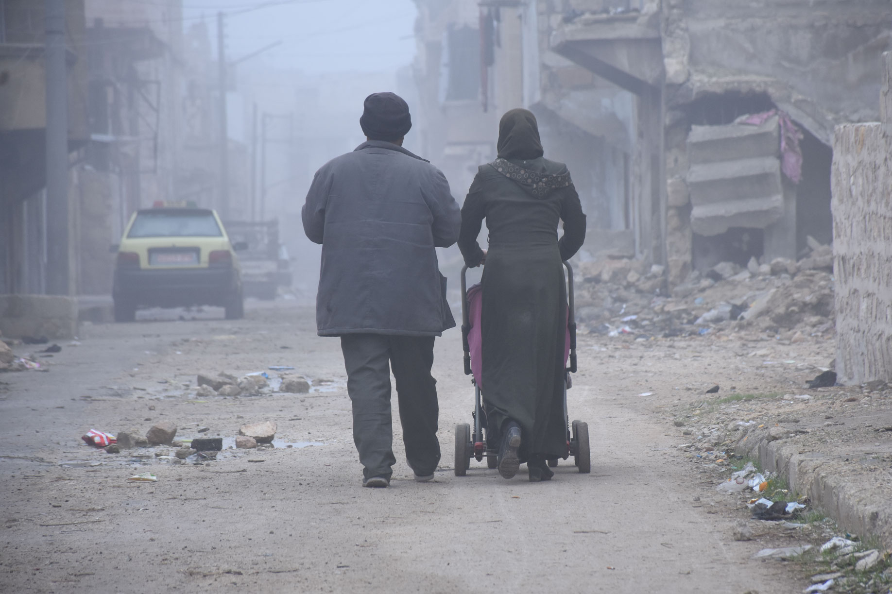 Parents walk their baby in a stroller in Aleppo. It is very cold and buildings around them are broken and destroyed.